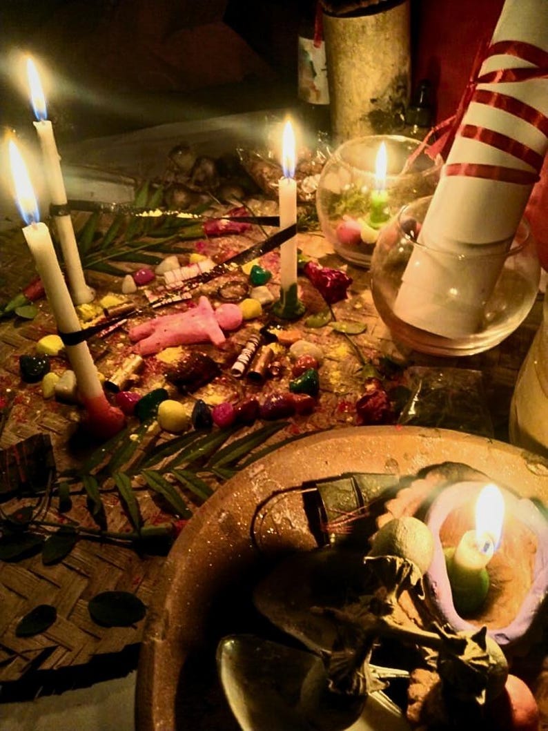 Puede incluir: Una colecci&oacute;n de objetos rituales que incluye varias velas blancas encendidas, una peque&ntilde;a figura rosa, piedras de colores, hojas verdes y un pergamino blanco enrollado con una cinta roja, dispuestos sobre una estera tejida con un cuenco de barro.