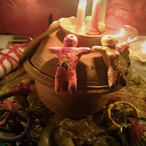 Puede incluir: Dos peque&ntilde;as figuras de arcilla rosa con forma humana y alfileres insertados est&aacute;n colocadas sobre la tapa de una vasija de cer&aacute;mica marr&oacute;n. Dos velas encendidas, una blanca y otra rosa, se encuentran en un cuenco sobre la tapa.