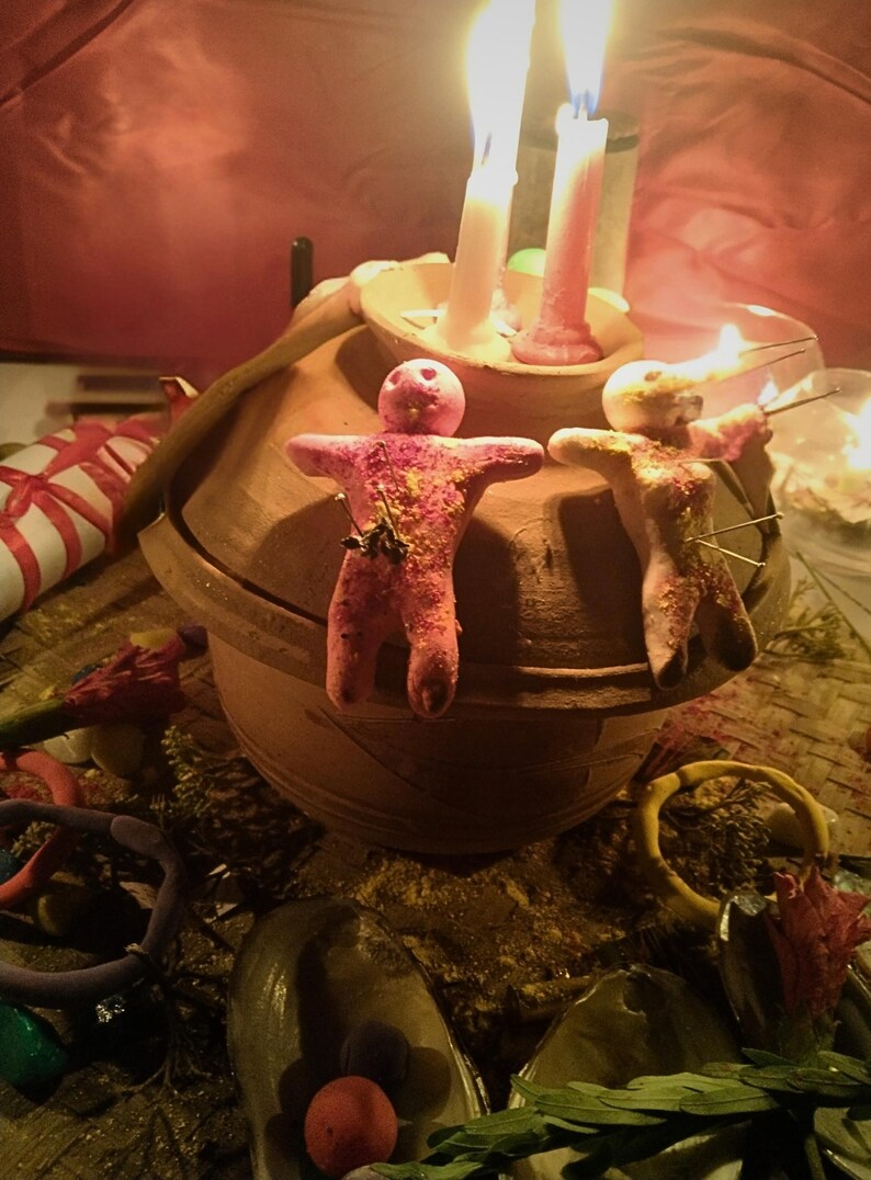 Puede incluir: Dos peque&ntilde;as figuras de arcilla rosa con forma humana y alfileres insertados est&aacute;n colocadas sobre la tapa de una vasija de cer&aacute;mica marr&oacute;n. Dos velas encendidas, una blanca y otra rosa, se encuentran en un cuenco sobre la tapa.