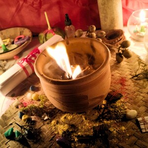 Puede incluir: Una vasija de barro con una llama encendida, rodeada de peque&ntilde;as piedras de colores esparcidas, materia vegetal seca, un pergamino enrollado atado con cinta roja, un frasco cuentagotas de vidrio y velas encendidas en un entorno tenue.