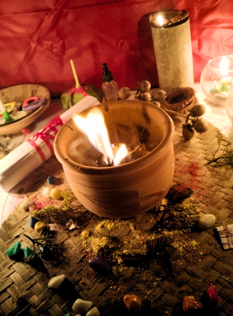Puede incluir: Una vasija de barro con una llama encendida, rodeada de peque&ntilde;as piedras de colores esparcidas, materia vegetal seca, un pergamino enrollado atado con cinta roja, un frasco cuentagotas de vidrio y velas encendidas en un entorno tenue.