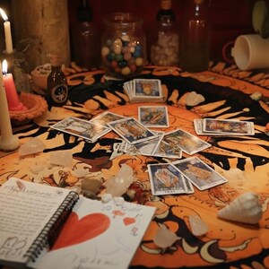 K&ouml;nnte beinhalten: Ein rundes orange-schwarzes Tuch mit Sonnendesign, bedeckt mit verstreuten Tarotkarten, brennenden Kerzen, klaren Kristallen, Muscheln und einem Spiralblock mit einem handgezeichneten roten Herzen und handschriftlichem Text in schwarzer Tinte.