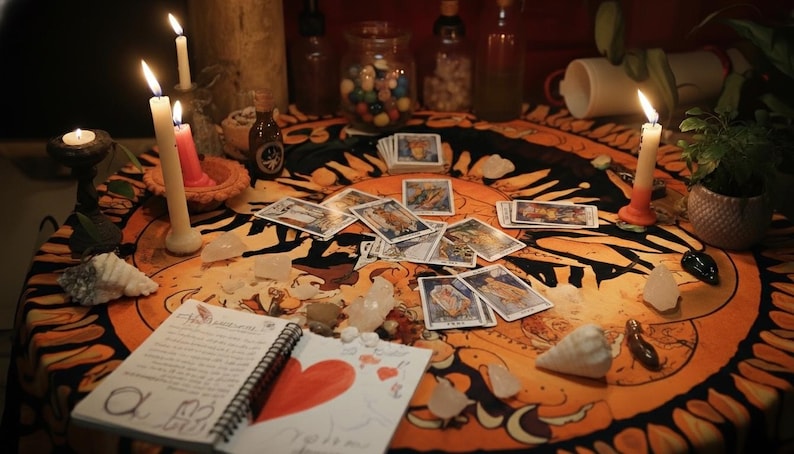 K&ouml;nnte beinhalten: Ein rundes orange-schwarzes Tuch mit Sonnendesign, bedeckt mit verstreuten Tarotkarten, brennenden Kerzen, klaren Kristallen, Muscheln und einem Spiralblock mit einem handgezeichneten roten Herzen und handschriftlichem Text in schwarzer Tinte.