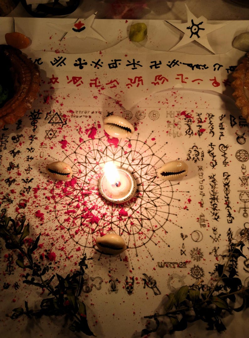 Puede incluir: Una hoja de papel blanca con un dise&ntilde;o de mandala geom&eacute;trico central, rodeado por cuatro conchas de cauri y una vela de t&eacute; encendida. El papel est&aacute; cubierto de s&iacute;mbolos de tinta negra y polvo rojo, con dos estrellas de papel arriba y follaje verde.