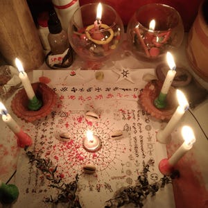 May include: A white paper sheet featuring intricate black geometric patterns, symbols, and text, surrounded by six lit white candles in clay holders, four cowrie shells, two glass bowls filled with colorful stones, and scattered red powder.