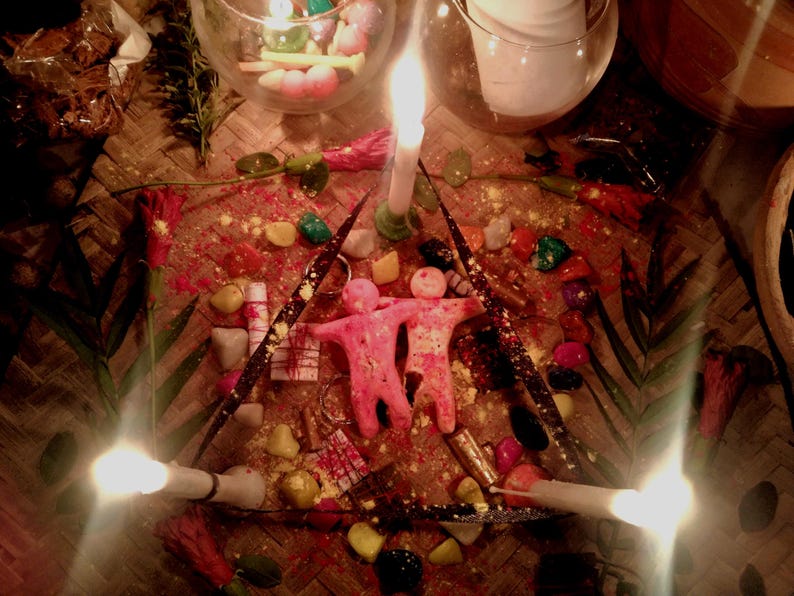 Puede incluir: Dos figuras de cera rosa se encuentran lado a lado dentro de un marco triangular formado por tres velas blancas encendidas. El arreglo est&aacute; rodeado de piedras coloridas dispersas, peque&ntilde;os paquetes envueltos y hojas verdes.