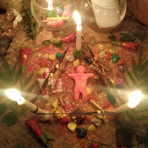 May include: A ritual arrangement featuring a central pink human-shaped wax figure surrounded by three lit white taper candles, colorful stones, red flowers, and small metallic objects on a woven mat, with two glass bowls containing candles and paper.