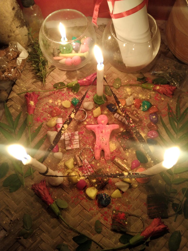 May include: A ritual arrangement featuring a central pink human-shaped wax figure surrounded by three lit white taper candles, colorful stones, red flowers, and small metallic objects on a woven mat, with two glass bowls containing candles and paper.