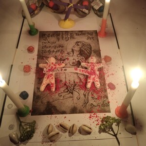 K&ouml;nnte beinhalten: Ein rituelles Arrangement mit einem zentralen Druck einer Figur mit Schriftzeichen, zwei kleinen wei&szlig;en menschen&auml;hnlichen Figuren, die durch einen Papierstreifen verbunden sind, vier brennenden Kerzen, rotem Pulver, Kaurimuscheln und einem violetten Kreuz.