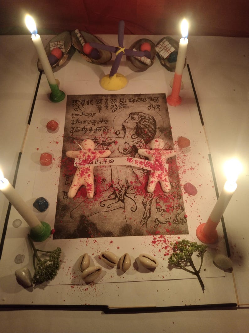 K&ouml;nnte beinhalten: Ein rituelles Arrangement mit einem zentralen Druck einer Figur mit Schriftzeichen, zwei kleinen wei&szlig;en menschen&auml;hnlichen Figuren, die durch einen Papierstreifen verbunden sind, vier brennenden Kerzen, rotem Pulver, Kaurimuscheln und einem violetten Kreuz.