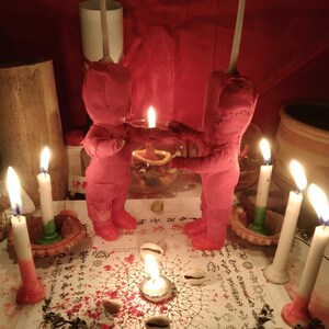 May include: Two pink fabric dolls stand facing each other on a white paper surface covered in black printed symbols and text. Each doll has a tall white candle attached to its head. Six additional white candles and several cowrie shells surround them.
