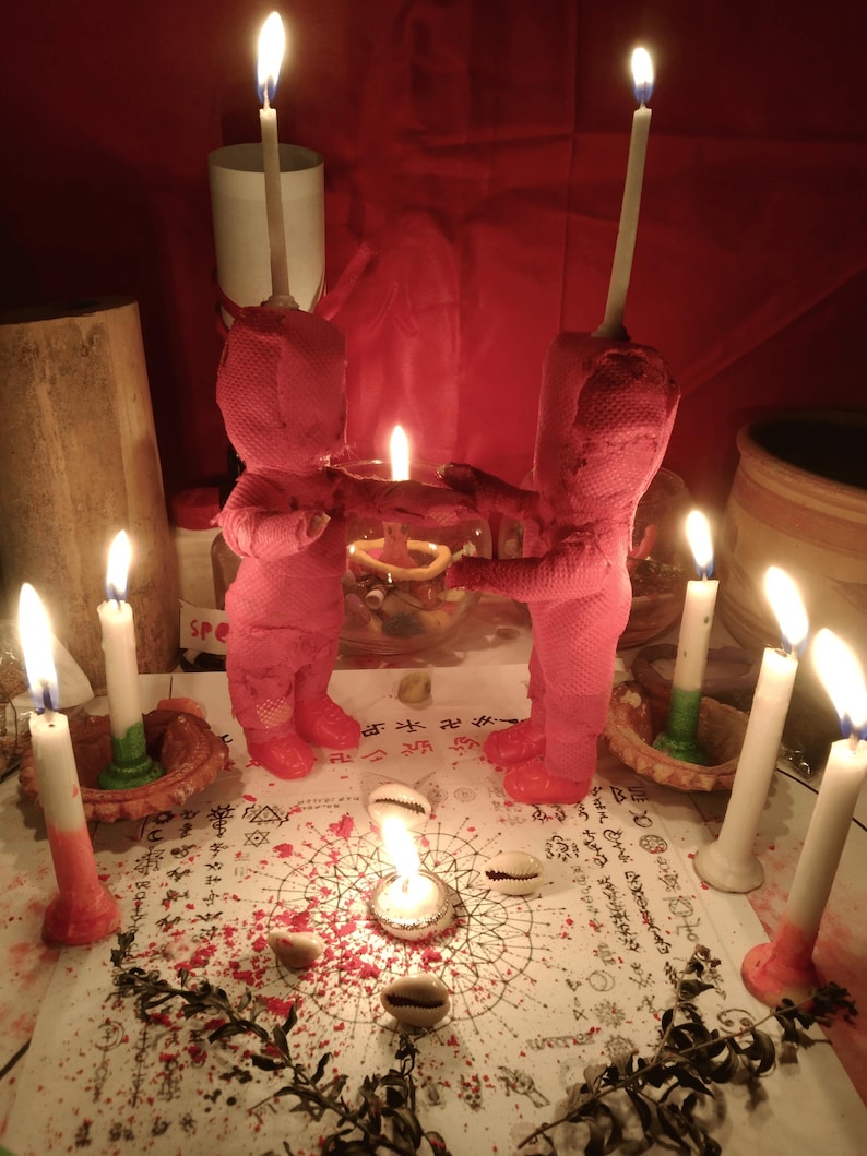 May include: Two pink fabric dolls stand facing each other on a white paper surface covered in black printed symbols and text. Each doll has a tall white candle attached to its head. Six additional white candles and several cowrie shells surround them.