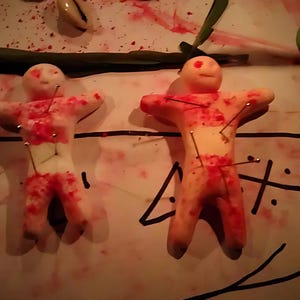 Puede incluir: Dos figuras de forma humana, de color claro y hechas de un material similar a la arcilla, cada una atravesada por varios alfileres met&aacute;licos. Las figuras presentan salpicaduras rojas y est&aacute;n colocadas sobre una superficie con marcas de tinta negra.