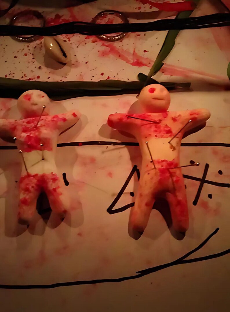 Puede incluir: Dos figuras de forma humana, de color claro y hechas de un material similar a la arcilla, cada una atravesada por varios alfileres met&aacute;licos. Las figuras presentan salpicaduras rojas y est&aacute;n colocadas sobre una superficie con marcas de tinta negra.
