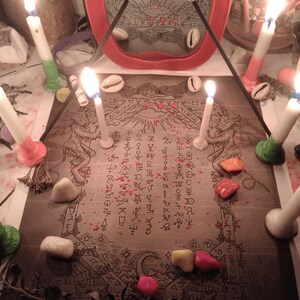 May include: A rectangular print featuring intricate line art of mythical creatures and occult symbols, surrounded by six lit white candles with colored bases, scattered cowrie shells, small colorful stones, and a red-framed mirror in the background.