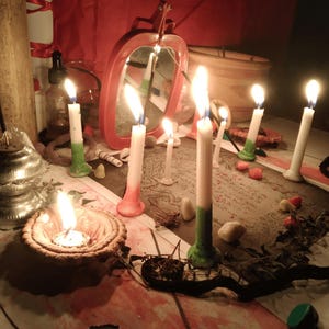 May include: A collection of lit white candles with colored bases, including green and red, arranged on a surface with a printed paper featuring text and illustrations. A red-framed mirror, a small clay bowl with a flame, and scattered stones are present.