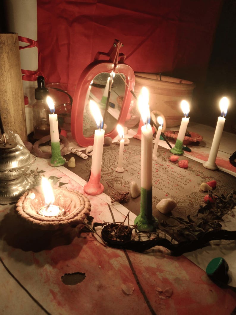 May include: A collection of lit white candles with colored bases, including green and red, arranged on a surface with a printed paper featuring text and illustrations. A red-framed mirror, a small clay bowl with a flame, and scattered stones are present.