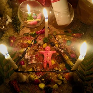 Puede incluir: Una figura de cera rosa con forma humana centrada dentro de un pentagrama de cinta negra, rodeada por cuatro velas blancas encendidas, piedras coloridas dispersas, peque&ntilde;os paquetes envueltos y p&eacute;talos de flores rojas sobre una estera tejida.