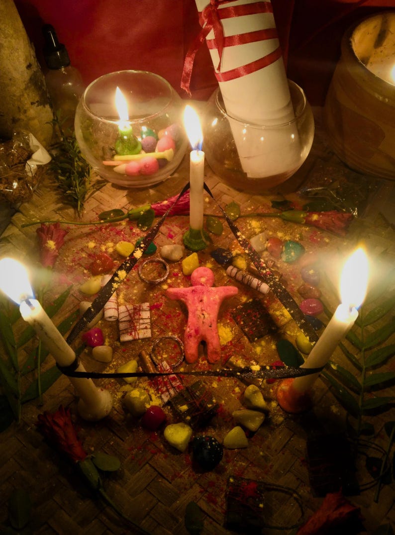 Puede incluir: Una figura de cera rosa con forma humana centrada dentro de un pentagrama de cinta negra, rodeada por cuatro velas blancas encendidas, piedras coloridas dispersas, peque&ntilde;os paquetes envueltos y p&eacute;talos de flores rojas sobre una estera tejida.