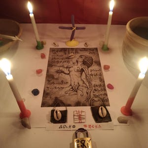 K&ouml;nnte beinhalten: Ein rituelles Arrangement mit vier brennenden Kerzen, einem zentralen Druck einer Figur mit komplexen Linien und Schriftzeichen, zwei kleinen dunklen Beuteln mit Kaurimuscheln, einem kleinen gewickelten B&uuml;ndel und einem Messingvorh&auml;ngeschloss.