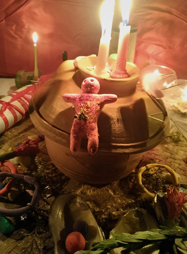 Puede incluir: Una vasija de barro con tapa, que presenta una peque&ntilde;a figura humanoide rosa adherida al frente. Dos velas encendidas est&aacute;n colocadas sobre la tapa. La escena incluye objetos dispersos como conchas, peque&ntilde;as esferas de colores y vegetaci&oacute;n.