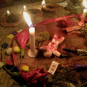 Puede incluir: Una efigie de arcilla blanca con marcas rojas y agujas, rodeada de velas blancas encendidas, peque&ntilde;as l&aacute;mparas de aceite de arcilla, piedras pulidas de colores, flores rojas secas y una etiqueta de papel con el texto V.F.T. sobre una estera.