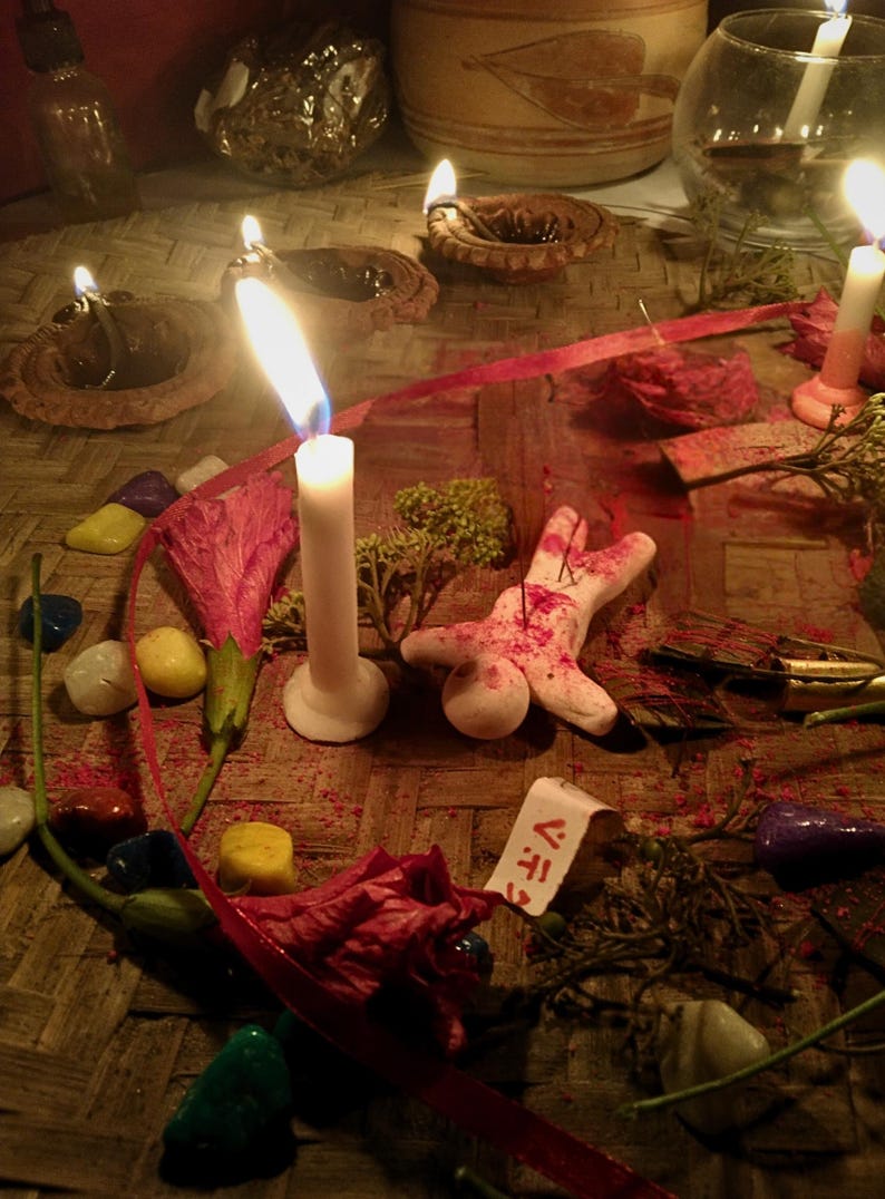 Puede incluir: Una efigie de arcilla blanca con marcas rojas y agujas, rodeada de velas blancas encendidas, peque&ntilde;as l&aacute;mparas de aceite de arcilla, piedras pulidas de colores, flores rojas secas y una etiqueta de papel con el texto V.F.T. sobre una estera.