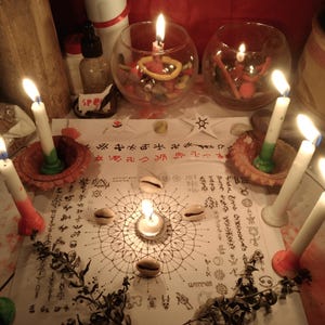 May include: A white paper sheet featuring intricate black geometric patterns, symbols, and text, surrounded by six lit white candles in clay holders and glass bowls. Four cowrie shells are placed on the paper, with dried plant sprigs nearby.