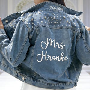 Puede incluir: Chaqueta vaquera azul claro adornada con perlas de imitación en los hombros. La parte trasera presenta el texto bordado "Mrs. Hranke" y la fecha "13.06.2026". Una chaqueta de novia elegante y personalizada.