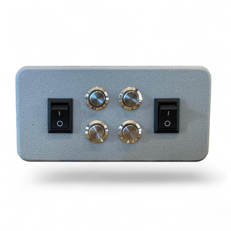 Puede incluir: Una caja de control rectangular gris con dos interruptores basculantes negros y cuatro botones plateados. Los interruptores est&aacute;n situados a la izquierda y a la derecha, mientras que los botones est&aacute;n en el centro.