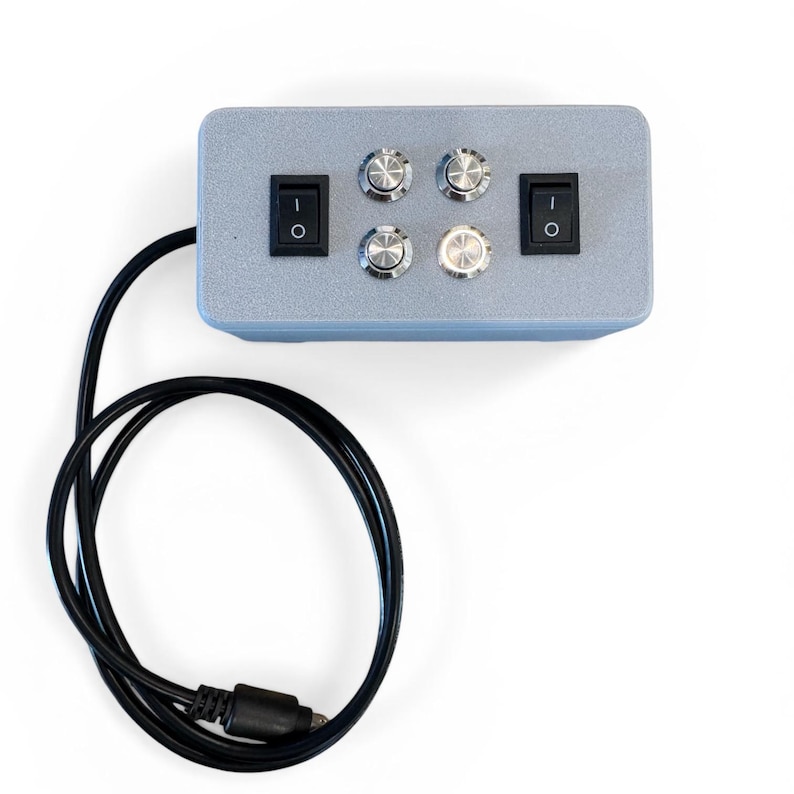 Puede incluir: Una caja de control gris con dos interruptores de encendido/apagado negros, cuatro perillas plateadas y un cable negro con un conector. La caja es rectangular y tiene un acabado mate. Las perillas tienen un dise&ntilde;o texturizado.
