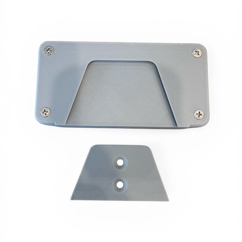 Puede incluir: Un soporte de pared gris claro impreso en 3D con un recorte trapezoidal y cuatro tornillos. Debajo, una pieza trapezoidal gris a juego con dos agujeros. Los art&iacute;culos est&aacute;n sobre un fondo blanco.