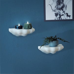 May include: Two white cloud-shaped shelves mounted on a blue wall. One shelf holds two candle holders with lit candles. The other shelf holds a small blue pot with a green plant. A framed black and white abstract art piece hangs above the shelves.