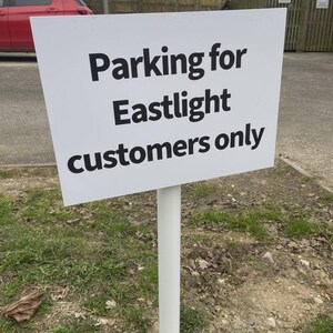 Könnte beinhalten: Ein weißes Schild mit schwarzem Text "Parken für Eastlight-Kunden nur" auf einem weißen Pfosten. Das Schild befindet sich in einem grasbewachsenen Bereich, mit einem roten Auto und Holzstrukturen im Hintergrund.