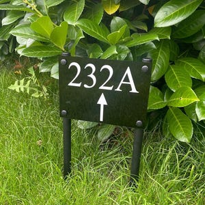 May include: A black address sign displaying the number 232A in white, with a white upward-pointing arrow. The sign is supported by two black posts and positioned in green grass, against a background of green foliage.