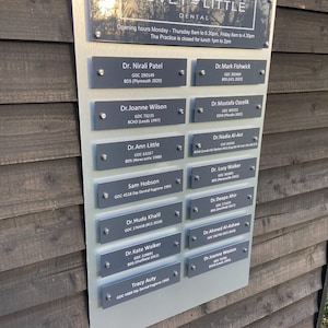 May include: A silver and clear sign for Tovey Little Dental, with the practice name and opening hours. Below are multiple nameplates with names and credentials of dental professionals.