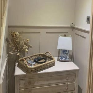 May include: A pale wooden chest of drawers with three drawers, a woven tray with a remote and headphones, and a lamp with a blue and white patterned base. Dried flowers in a vase are on the chest. The room has panelled walls.