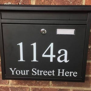 May include: Black mailbox with the address "114a" and the text "Your Street Here" in white. The mailbox has a lock and a small slot for mail. The mailbox is mounted on a brick wall.