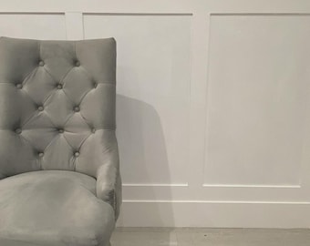 Puede incluir: Una silla gris tapizada en terciopelo con botones y reposabrazos. La silla está colocada contra una pared blanca con paneles y un suelo de madera clara. La silla tiene patas de madera clara.