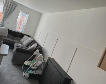 Puede incluir: Un salón con una alfombra gris, un sofá gris y una silla de bebé. Una cubierta de radiador negra está en primer plano. Las paredes son blancas con paneles decorativos. Se ve una ventana con cortinas grises.
