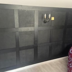 May include: A dark grey panelled wall with a decorative sconce and a portion of a bed. The wall is divided into rectangular sections. The sconce has two light bulbs. The bed has a blue headboard and a red patterned duvet.