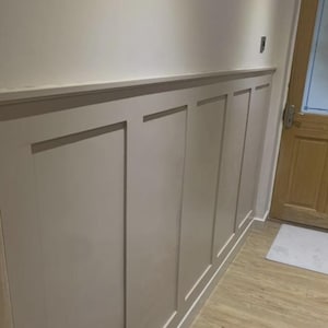 May include: A section of a wall with light beige wainscoting. The wainscoting is divided into rectangular panels with a simple, clean design. The wall is painted in a neutral color, and the floor is a light wood.