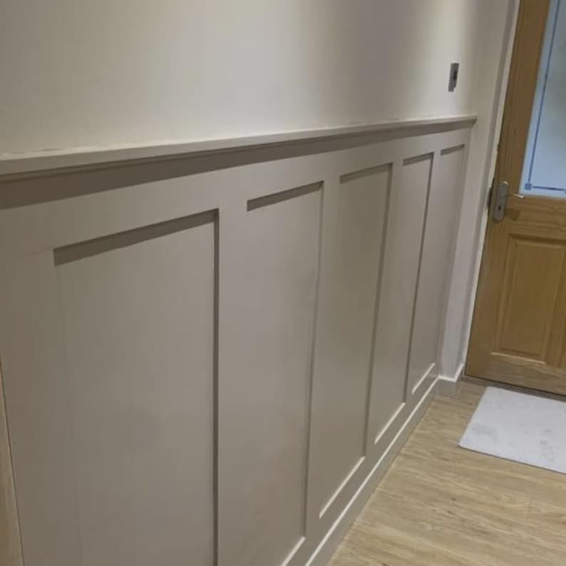 May include: A section of a wall with light beige wainscoting. The wainscoting is divided into rectangular panels with a simple, clean design. The wall is painted in a neutral color, and the floor is a light wood.