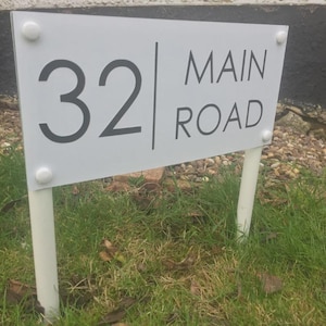 May include: A white rectangular sign displaying the number "32" and the words "MAIN ROAD" in black. The sign is attached to two white posts with white fixings. The sign is positioned in a grassy area.