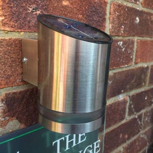 May include: A cylindrical, brushed-metal outdoor solar light fixture mounted on a brick wall. The light has a dark, angled top and a clear bottom section. The fixture is attached to a rectangular metal bracket.