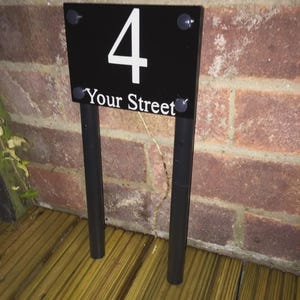 May include: A black street sign displaying the number "4" in white and the words "Your Street". The sign is supported by two black posts. The background is a red brick wall.