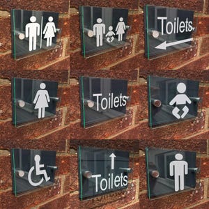 May include: A set of clear glass toilet signs featuring white icons of male, female, family, and disabled symbols, plus the word "Toilets". The signs are mounted on a brick wall with silver fixings.