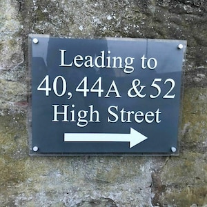 Könnte beinhalten: Rechteckiges Schild mit der Aufschrift "Leading to 40,44A & 52 High Street" und einem weißen Pfeil nach rechts. An einer Steinmauer befestigt.