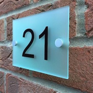 May include: A rectangular, frosted glass house number sign with the number "21" in black. The sign is mounted on a brick wall with two silver-colored fasteners.