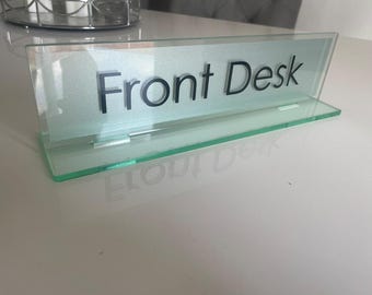 May include: A clear glass sign displaying the words "Front Desk" in black. The sign has a light green tint and a matching base. It is designed to stand on a desk or counter, providing clear information.